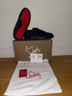 Svarta sneakers från Christian Louboutin - Snygga svarta sneakers från Christian Louboutin med klassisk röd sula och diskreta nitar på ovandelen. Skorna är låga och har en lyxig känsla med detaljer i mocka och läder. Perfekta för dig som vill sticka ut med exklusiv stil.