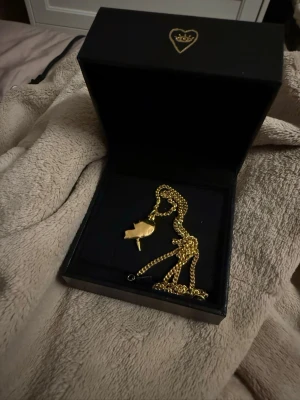 Guldigt Maria Nilsdotter  halsband med hjärthänge - Snyggt halsband i guldfärgad metall med ett hjärtformat hänge. Kedjan är tunn och elegant, och hänget har en stilren, blank finish. Kommer i en svart ask med hjärtdetalj på locket. Perfekt accessoar för att lyfta din outfit.