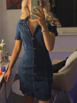 Blå jeansklänning med knappar från Zara - Snygg blå jeansklänning med korta ärmar och knappar hela vägen framtill. Klänningen har en figurnära passform och klassisk krage, perfekt för en trendig och avslappnad look.