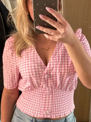 Rutig rosa blus med v-ringning - Supersöt rosa och vit rutig blus med v-ringning och knappar framtill. Blusen har korta ärmar och smockad midja som ger en snygg passform. Perfekt till jeans eller kjol för en trendig look.