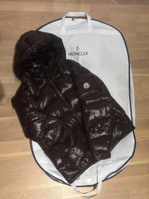 Moncler Maya - brun  - Säljer min fina och unika bruna Moncler maya - Dustbag medföljer - Ny modell där badgen inte skannar - Allting stämmer i överens - Lite små repor på dragkedjan men annars inga skador - Kom med bud!