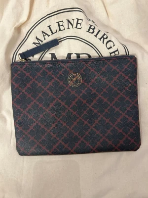 Purse från By Malene Birger - Snygg purse från By Malene Birger i svart och vinrött med klassiskt mönster och guldfärgad logga framtill. Väskan har dragkedja upptill och är gjord i tåligt konstläder. Perfekt för att bära vardagliga artiklar i. OBS: dustbagen i bakgrunden ingår ej i köp!