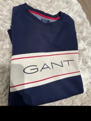 Marinblå sweatshirt från GANT - Snygg marinblå sweatshirt från GANT med vit panel och röda detaljer över bröstet. Stor GANT-logga framtill. Klassisk rund halsringning och långa ärmar. Perfekt för en avslappnad och stilren look.