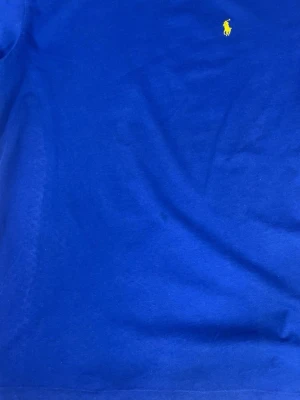 Blå t-shirt från Ralph Lauren - Klassisk blå t-shirt från Ralph Lauren med rund halsringning och den ikoniska gula logobroderingen på bröstet. Tillverkad i mjuk bomull som känns skön mot huden. Perfekt för dig som gillar stilrena och tidlösa plagg.