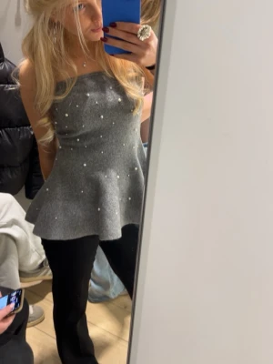 Grå axelbandslös topp med pärlor - Säljer en grå axelbandslös topp med utsvängd peplumform och små pärlor som dekoration. Toppen har en bandeau-liknande överdel och är perfekt för dig som vill ha något stilrent men ändå unikt. Passar snyggt till både jeans och byxor.