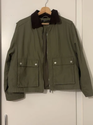 Olivgrön jacka med brun krage - Snygg olivgrön jacka från Gina  med kontrasterande brun krage. Jackan har två stora fickor framtill med tryckknappar och dragkedja hela vägen upp. Perfekt för dig som gillar minimalistisk och clean stil.