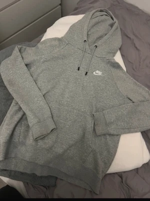 Grå hoodie från Nike - Klassisk grå hoodie från Nike med vit broderad logga på bröstet. Tröjan har huva med dragsko, känguruficka och ribbade muddar vid ärmslut och nederkant. Perfekt för en avslappnad och sportig stil.