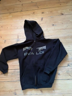 Svart hoodie med dragkedja och tryck - Svart hoodie från Bvaldi med dragkedja framtill och två fickor. På bröstet finns ett tryck med två händer och texten 'BVALDI' i rhinestones. Hoodien har ribbade muddar vid ärmslut och nederkant samt en klassisk huva. Perfekt för en avslappnad streetwear-look. Skriv om det är något. Priset går såklart att diskutera.