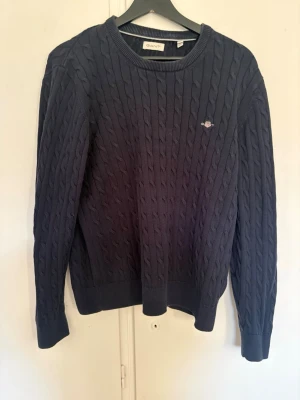 Mörkblå kabelstickad tröja GANT - Kabelstickad tröja i mörkblått från GANT med rund halsringning och diskret logga broderad på bröstet. Tröjan har långa ärmar, ribbade muddar och klassisk passform. Perfekt för lager på lager och en stilren look.