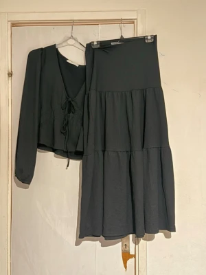 Svart set med långärmad topp och kjol - Snyggt svart set med långärmad topp som knyts framtill och en matchande lång kjol med volanger. Setet har en mjuk och luftig känsla, perfekt för dig som gillar en enkel men stilren look. Toppen är L och kjolen S 