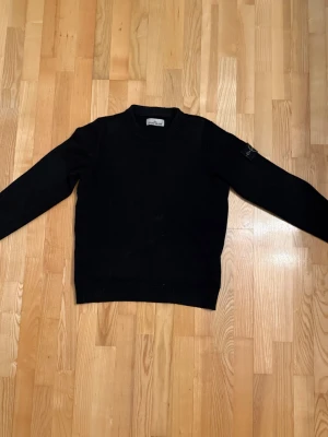 Svart stickad tröja från Stone Island - Snygg svart stickad tröja från Stone Island med klassisk rund hals och lång ärm. Tröjan har den ikoniska Stone Island-loggan på ärmen och är tillverkad i mjukt material som passar perfekt till höst och vinter. Använt ett par gånger, digitalt kvitto finns med, köpt på careofcarl