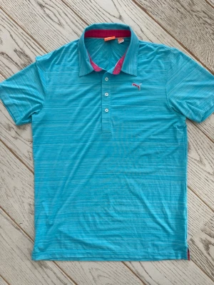 Turkos pikétröja från Puma - Snygg turkos pikétröja från Puma med diskreta ränder och rosa detaljer på insidan av kragen. Tröjan har korta ärmar, knappslå med fyra knappar och Puma-logga på bröstet. Perfekt för en sportig och avslappnad stil.