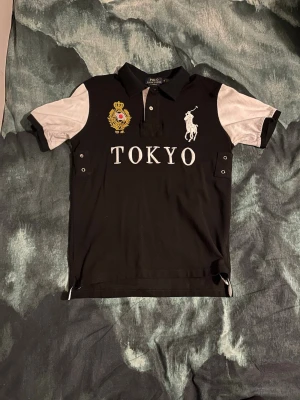 Polo Ralph Lauren Tokyo - Snygg svart pikétröja från Polo Ralph Lauren med vita ärmar och detaljer. Stort Tokyo-tryck på bröstet, broderad logga och emblem. Klassisk krage och knappar framtill. Perfekt för dig som gillar streetstyle och sportig look.