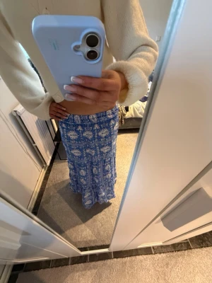 Blå blommig långkjol - Säljer en blå långkjol med vitt blommigt mönster. Kjolen har en avslappnad passform och är perfekt för varma dagar. Materialet känns lätt och flowy, vilket gör den superbekväm att bära. Passar perfekt till en enkel topp eller stickad tröja.