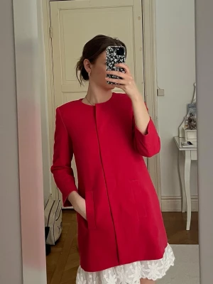 Röd minimalistisk kavaj - Stilren röd kavaj med rak passform och dold knäppning framtill. Modellen har trekvartsärmar och två diskreta fickor. Kappan är enkel och modern. 