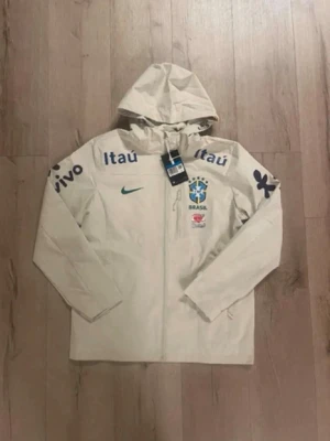 Nike Brasil vindjacka med huva - Stilren beige vindjacka från Nike med Brasiliens landslagslogga och sponsorer tryckta på bröst och ärmar. Jackan har huva, hel dragkedja och långa ärmar. Perfekt för dig som gillar sportig stil och vill representera Brasilien.