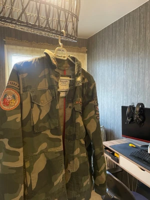 Camo overshirt från Abercrombie & Fitch - Snygg camouflagefärgad overshirt från Abercrombie & Fitch med coola patchar på ärmen och bröstet. Jackan har flera fickor framtill, knappstängning och detaljer i rött. Tillverkad i slitstarkt bomullsmaterial och har en avslappnad passform.