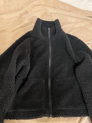 Svart teddyjacka med dragkedja - Mysig svart teddyjacka med hög krage och hel dragkedja framtill. Jackan har två stora fickor och är gjord i fluffigt, mjukt material som håller dig varm. Perfekt för kyliga dagar och enkel att matcha med olika outfits. Den har ett litet slit på sidan av ena  fickan, det var sömnen som löstes upp lite men går enkelt att sy ihop hemma själv. Skriv gärna till mig vid mer frågor/bilder