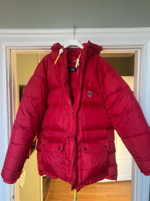 Röd dunjacka från Fjällräven - Riktigt fet röd dunjacka från Fjällräven med huva och gula detaljer på snörena. Jackan har två stora fickor framtill med knappar och dragkedja, samt Fjällräven-logga på bröstet. Perfekt för kalla vinterdagar och riktigt varm.