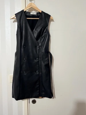 Svart omlottklänning i skinnimitation - Snygg svart omlottklänning från Mango i skinnimitation med dubbelknäppning framtill och bälte i midjan. Ärmlös design och kort längd ger en modern look.