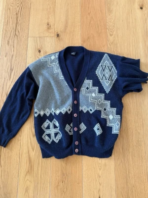 Nische Mörkblå kofta  - Snygg mörkblå kofta thriftad i Frankrike med grå och vita geometriska mönster och bruna knappar. Koftan har v-ringning och ribbade muddar. Perfekt för dig som gillar retrostil och vill sticka ut lite extra.