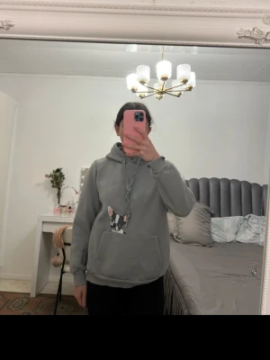 Grå hoodie med hundtryck - Säljer en grå hoodie med stor ficka framtill och en söt hundillustration på magen. Hoodien har huva med dragsko och ribbade muddar vid ärmslut och nederkant. Perfekt för dig som gillar djurmotiv och vill ha en chill stil.