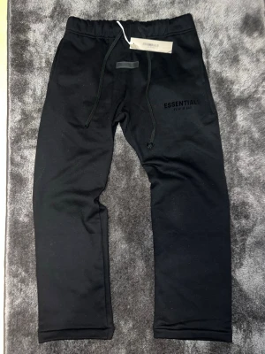 Svarta Essentials mjukisbyxor - Säljer ett par svarta mjukisbyxor från Fear of God Essentials med dragsko i midjan och logga tryckt på vänster lår. Byxorna har en avslappnad passform och är tillverkade i mjukt material som passar perfekt för chill dagar. Stilsäkra och trendiga mjukisar!