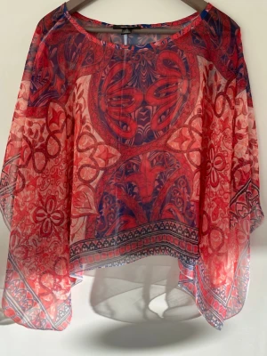 Färgglad topp - Ett riktigt unikt plagg i otroliga färger mellan blå och röd. Riktigt tunt tyg så passar bra över linne eller bh, men har stora armar så går säkerligen att addera som lager över en långarmad enfärgad tröja också. Storlek M men mycket stor och fungerar nog upp till xl 