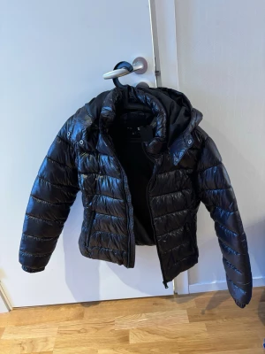 Svart pufferjacka med huva från lager157 - Snygg svart pufferjacka från lager157 med glansig finish och avtagbar huva. Jackan har dragkedja framtill, två sidofickor och quiltad design som ger en modern look. Perfekt för kalla vinterdagar och riktigt varm. Aldrig använd lappen kvar storlek M 