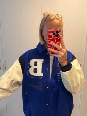 Blå och vit bomberjacka med brodyr - Snygg blå bomberjacka med vita ärmar och svarta detaljer. Stor broderad bokstav B på bröstet och baseboll-inspirerat broderi. Jackan har tryckknappar och klassisk collegekrage. Perfekt för en sportig och trendig look.