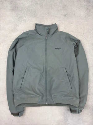 Grå vindjacka från Woolrich - Stilren grå vindjacka från Woolrich med hög krage och dragkedja framtill. Jackan har två snedställda fickor, ribbade muddar vid ärmslut och nederkant samt diskret logga på bröstet. Materialet är lätt och vindtätt, perfekt för blåsiga dagar.