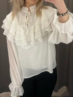 Vit volangblus med knyt - Säljer en romantisk vit blus med flera lager volanger över axlar och bröst, samt knytband vid halsen. Blusen är tillverkad i ett tunt, transparent tyg och har långa puffärmar med resår vid ärmslut. Perfekt för dig som gillar en feminin och trendig stil.