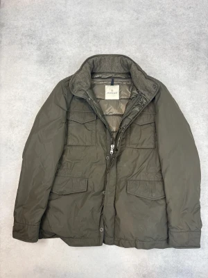 Olivgrön dunjacka från Moncler - Snygg olivgrön dunjacka från Moncler med fyra stora fickor framtill och hög krage med mjukt innerfoder. Jackan har dragkedja och knappar samt quiltad insida för extra värme. Perfekt för kalla vinterdagar och riktigt stilren look.