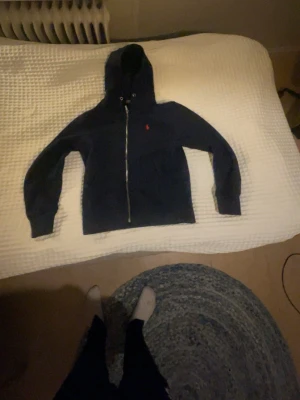 Mörkblå hoodie från Polo Ralph Lauren - Mörkblå hoodie med dragkedja från Polo Ralph Lauren. Klassisk design med huva och två fickor framtill. Ikoniska röda Polo-loggan broderad på bröstet. Tillverkad i mjukt bomullsmaterial som känns skönt mot huden. Perfekt för en avslappnad och stilren look.