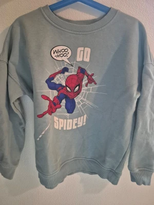 Barn Blå Spiderman sweatshirt stl 134 - Cool blå sweatshirt med Spiderman-tryck och texten 'GO SPIDEY!'. Tröjan har rund hals och långa ärmar, perfekt för dig som gillar Marvel och vill sticka ut med en färgglad och bekväm tröja.