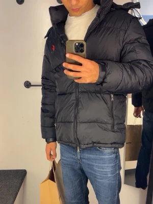Svart dunjacka från Polo Ralph Lauren - Snygg svart dunjacka från Polo Ralph Lauren med röd logga på bröstet. Jackan har huva, dragkedja framtill och två sidofickor med dragkedja. Perfekt för kalla vinterdagar och riktigt skön att bära.