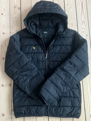 Svart pufferjacka Lyle & Scott - Svart pufferjacka från Lyle & Scott Junior med huva och dragkedja framtill. Jackan har det klassiska gula logomärket på bröstet och är quiltad för extra värme. Perfekt för kalla dagar och enkel att matcha med olika outfits.