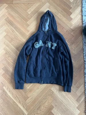 Mörkgrå hoodie med dragkedja från GANT - En snygg mörkgrå ziphoodie från GANT i storlek L. Loggan är snyggt broderat på framsidan och tröjan är i mycket bra skick.