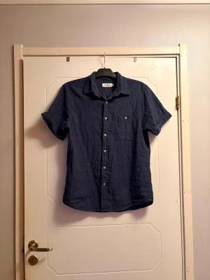 Mörkblå linneskjorta kort ärm - Snygg mörkblå skjorta i linne med korta ärmar och klassisk krage. Skjortan har knappar framtill och en bröstficka. Perfekt för en avslappnad och stilren look.