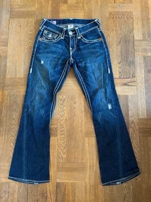 True Religion bootcut jeans blå - Säljer ett par blå bootcut jeans från True Religion med tydliga kontrastsömmar och slitna detaljer. Jeansen har fem fickor, dekorativa bakfickor med lock och knappar samt låg midja. Materialet är klassisk denim i bomull.