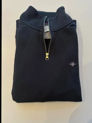 Marinblå halvzip från Gant - Helt ny Klassisk svart sweatshirt från Gant med halv dragkedja och liten broderad logga på bröstet. Tröjan har hög krage och är tillverkad i mjukt bomullsmaterial. Perfekt för en avslappnad och stilren look.