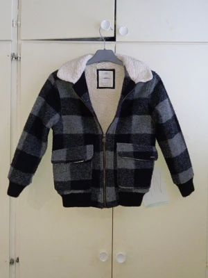 Rutig teddyjacka med dragkedja - Mysig teddyjacka med stora rutiga mönster i svart och grått. Jackan har en fluffig vit krage, två stora fickor framtill och dragkedja. På ryggen står det 'SOUND OF SEATTLE' broderat. Perfekt för kyliga dagar och riktigt snygg streetstil.