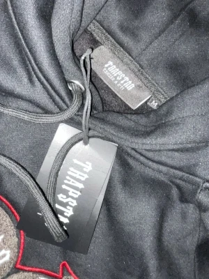 Svart Trapstar hoodie med tryck - Snygg svart hoodie från Trapstar med stort broderat tryck på bröstet där det står 'Shooter' i vitt och rött. Hoodien har huva med dragsko och ribbade muddar. Perfekt för dig som gillar streetwear och vill sticka ut med en fet logga framtill.