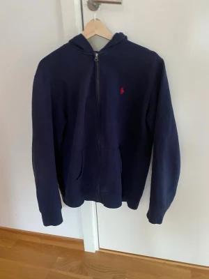 Mörkblå hoodie från Ralph Lauren - Mörkblå hoodie med dragkedja från Ralph Lauren. Klassisk design med röd broderad logga på bröstet, kängurufickor och huva. Tillverkad i mjukt material som passar perfekt för en avslappnad stil. Inga skador eller fläckar priset kan diskuteras 