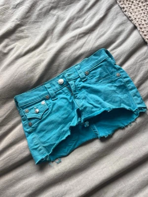 Turkos  jeanskjol  från True Religion - Säljer en turkos jeanskjol från True Religion med råa kanter och klassiska fem fickor. Kjolen har låg midja, silverfärgade knappar och nitar samt bälteshällor. Perfekta för varma dagar och en avslappnad stil.