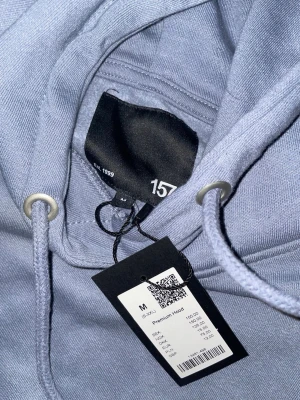 Ljusblå hoodie från 157 - Snygg ljusblå hoodie från 157 med klassisk huva och dragsko. Tillverkad i mjukt material och har en stor magficka framtill. Perfekt för en chill och avslappnad stil. Ribbstickade muddar vid ärmslut och nederkant för extra komfort. Priset går bra och diskutera 