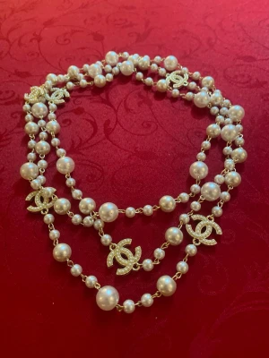 Chanel pärlhalsband med loggor - Lyxigt halsband från Chanel med vita pärlor och flera glittriga Chanel-loggor i guld. Halsbandet är långt och kan bäras i flera lager. Perfekt statement-piece för dig som vill sticka ut med exklusiva accessoarer.