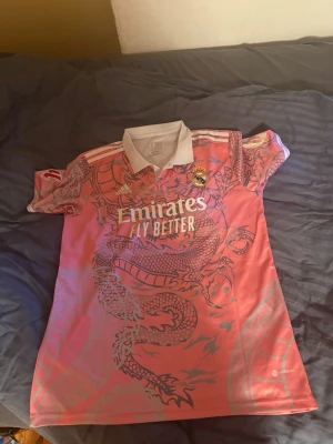 Rosa Real Madrid matchtröja drake - Säljer en rosa Real Madrid matchtröja från Adidas med stort drakmotiv i grått. Tröjan har vit krage, vita detaljer och tryck med 'Emirates Fly Better' på bröstet. På ryggen står det 'Mbappé' och nummer 9. Perfekt för fotbollsfans! Storlek är xl men är lite för sin storlek. Den passar mig perfekt som har m/l