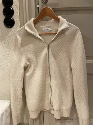 Beige cardigan stickad - Säljer en beige ribbad ziptröja från East West. Tröjan har krage, dragkedja framtill och är tillverkad i ett mjukt, stickat material. Perfekt för lager på lager och enkel att matcha med olika outfits.