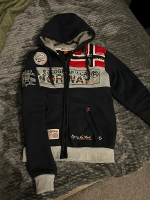Geographical Norway hoodie med tryck - Mörkblå hoodie från Geographical Norway med dragkedja, gråa muddar och detaljer i rött, vitt och grått. Stor logga och patchar på bröstet och ärmen, samt norsk flagga på axeln. Mjuk insida och fickor framtill. Perfekt för dig som gillar streetwear och snygga detaljer.